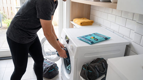 Iemand stopt donkere sportkleding in de wasmachine met een sporttas ernaast en een plat pak Floretti Fris Linnen wasstrips op de machine